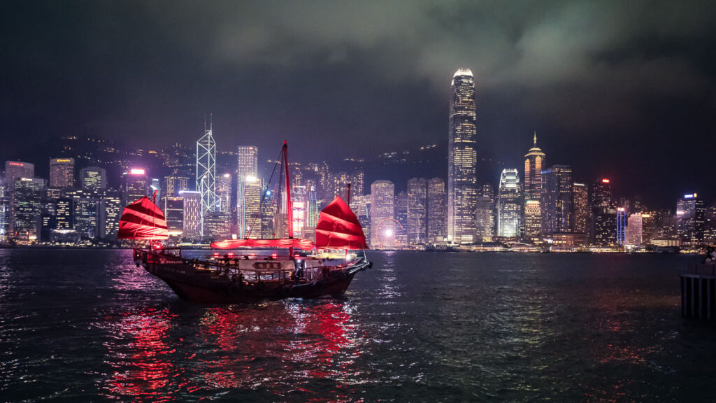 Traditionelle rote chinesische Dschunke (Aqua Luna) auf dem Victoria Harbour vor der beleuchteten Skyline von Hongkong Island bei Nacht.