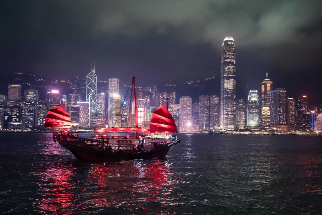 Traditionelle Dschunke mit roten Segeln im Victoria Harbour vor der beleuchteten Skyline – Perfekt für Nachtaufnahmen auch während der Regenzeit in Hongkong.