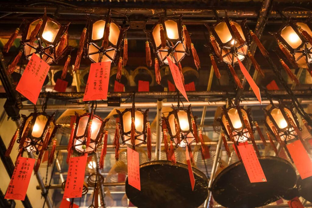 Traditionelle chinesische Laternen aus Holz mit warmem Licht und herabhängenden roten Wunschzetteln an der Decke eines Tempels in Hongkong.