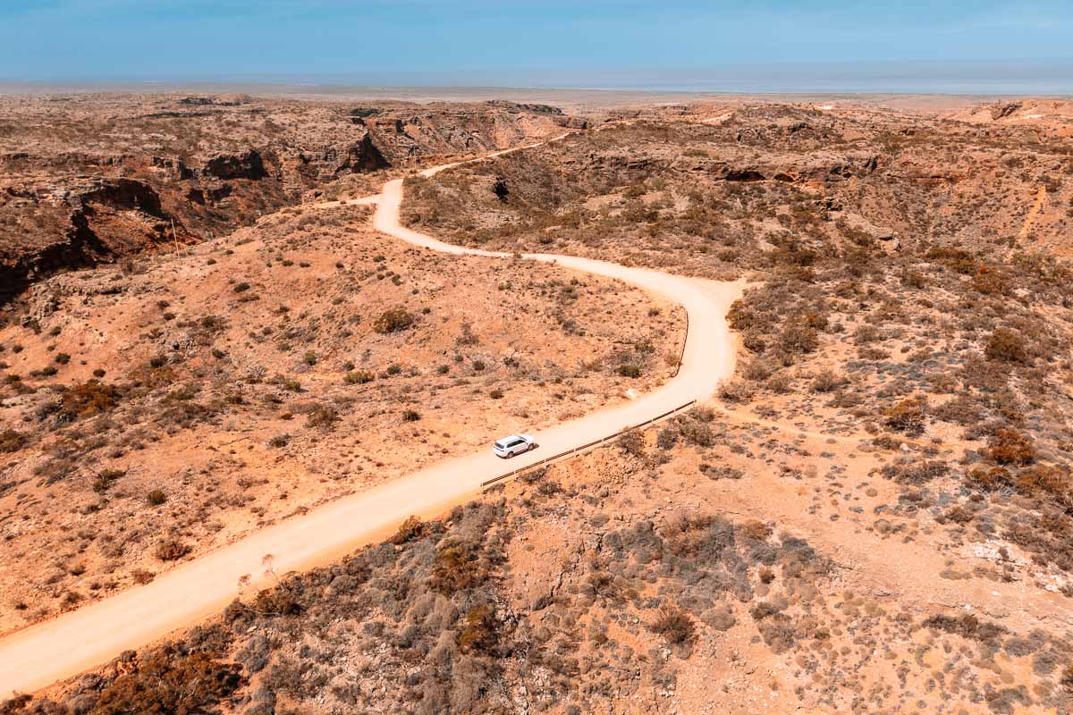 Drohnenaufnahme vom Charles Knife Canyon bei Exmouth in Westaustralien mit einer kurvigen Straße durch die rote Outback-Landschaft und einem weißen Auto.