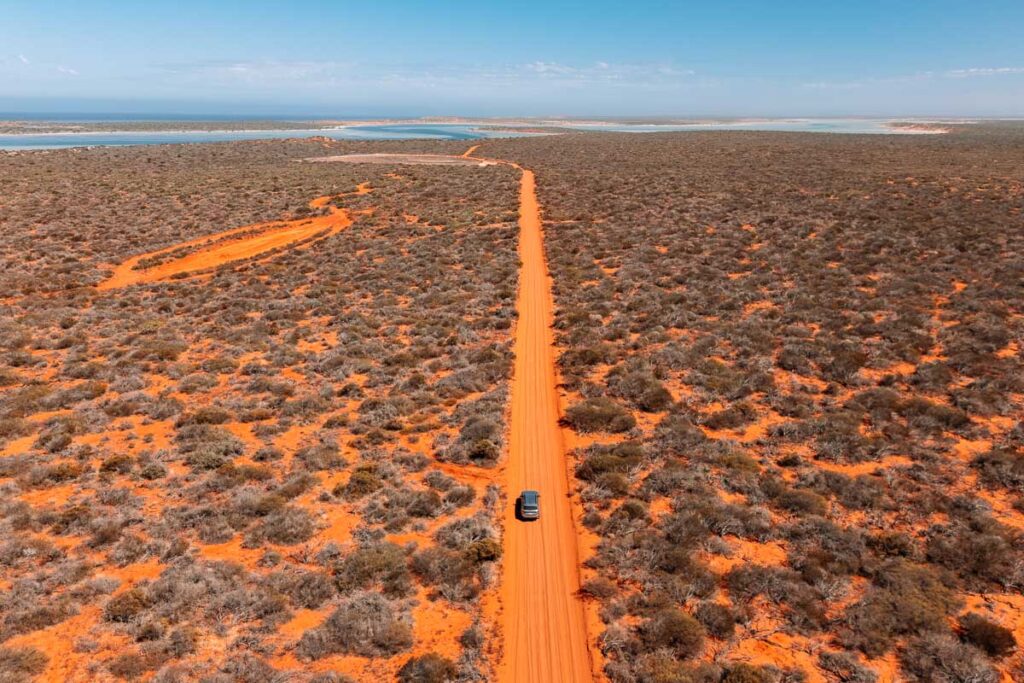 Drohnenaufnahme des Ford Everest von Auszeit Abenteuer auf einer schnurgeraden, leuchtend roten Sandpiste im Francois Peron Nationalpark. Das silberne Fahrzeug fährt durch eine weite, flache Landschaft mit dichtem, niedrigem Buschwerk auf roter Erde. Im Hintergrund ist eine blaue Lagunen- und Küstenlandschaft unter einem klaren Himmel zu sehen.