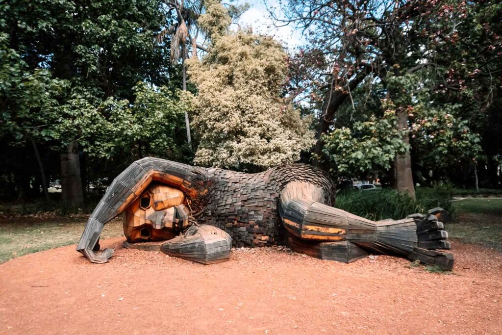 Hölzerne Skulptur der Giants of Mandurah, die entspannt auf der Seite auf dem Waldboden liegt. Kunstprojekt von Thomas Dambo in Westaustralien, umgeben von Bäumen.
