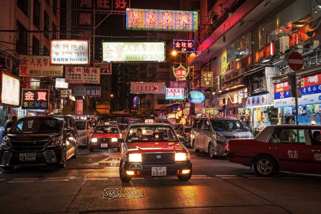 Rote Taxis im dichten Stadtverkehr von Hongkong bei Nacht unter bunten Neonreklamen – Stimmungsvolle Straßenszene während der Regenzeit in Hongkong.