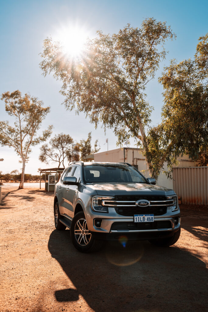 Ford Everest 4x4 Fahrzeug auf roter Erde, im Hintergrund Bäume, durch die sich die Sonne sternförmig bricht. Roadtrip Westaustralien, Billabong Roadhouse, Auszeit Abenteuer