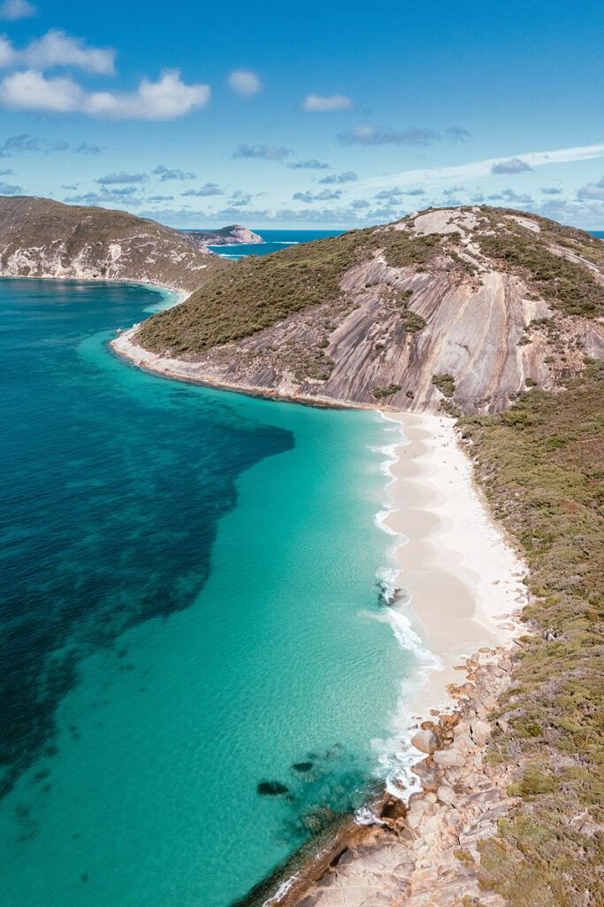 Atemberaubende Drohnenaufnahme vom Misery Beach im Torndirrup National Park, Westaustralien. Die Luftbildperspektive zeigt die geschützte Bucht mit ihrem strahlend weißen Sandstrand, der von massiven Granitfelsen und steilen, grünen Hängen eingerahmt wird. Das Wasser leuchtet in intensiven Türkis- und Blautönen und ist kristallklar. Im Hintergrund erstreckt sich der Ozean bis zum Horizont unter einem blauen Himmel.