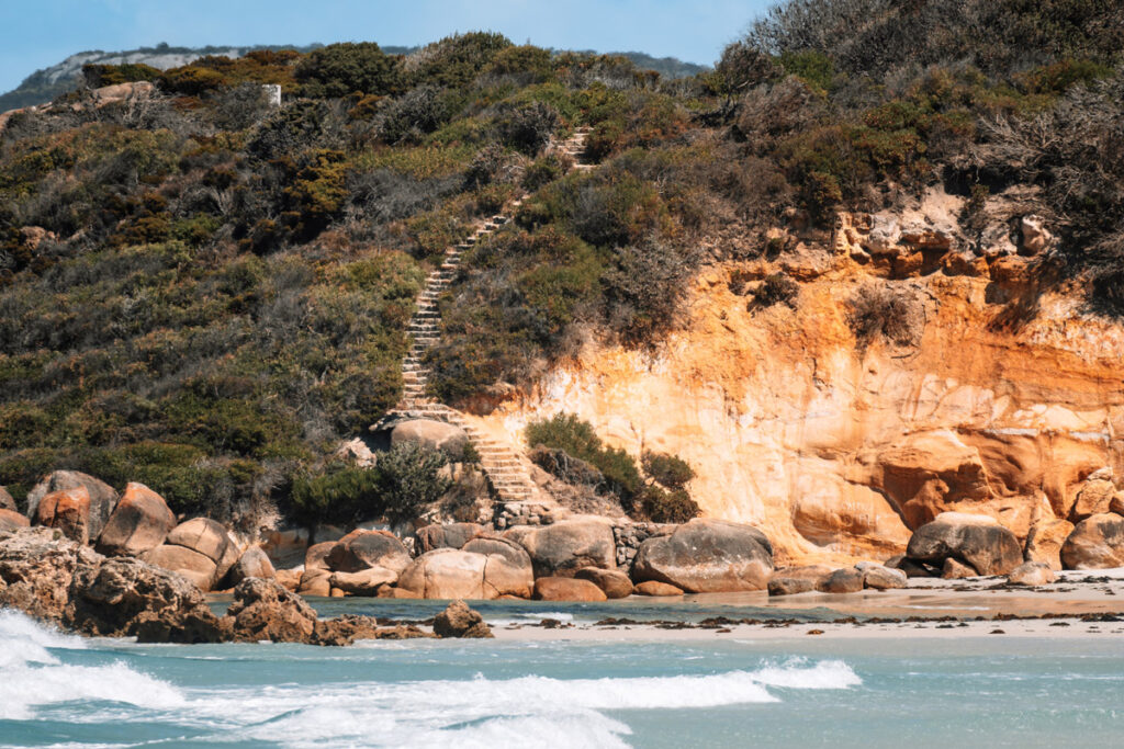 Malerische Küstenlandschaft am Nanarup Beach, Westaustralien. Eine steile Holztreppe führt einen bewachsenen Hang hinunter zum Strand. Die Klippen bestehen aus rötlichem Gestein, das einen schönen Kontrast zum grünen Gebüsch und dem weißen Sandstrand bildet. Im Vordergrund branden sanfte Wellen an die Felsen am Ufer.