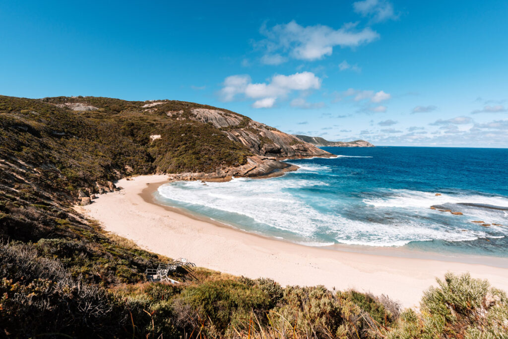 Malerische Bucht am Salmon Holes Beach im Torndirrup National Park, Westaustralien. Ein breiter, weißer Sandstrand schmiegt sich an steile, buschbewachsene Hänge. Kräftige Wellen des tiefblauen Ozeans rollen in die Bucht und brechen schäumend am Ufer. Im Vordergrund führt eine kleine Holztreppe durch die Vegetation hinunter zum Strand.