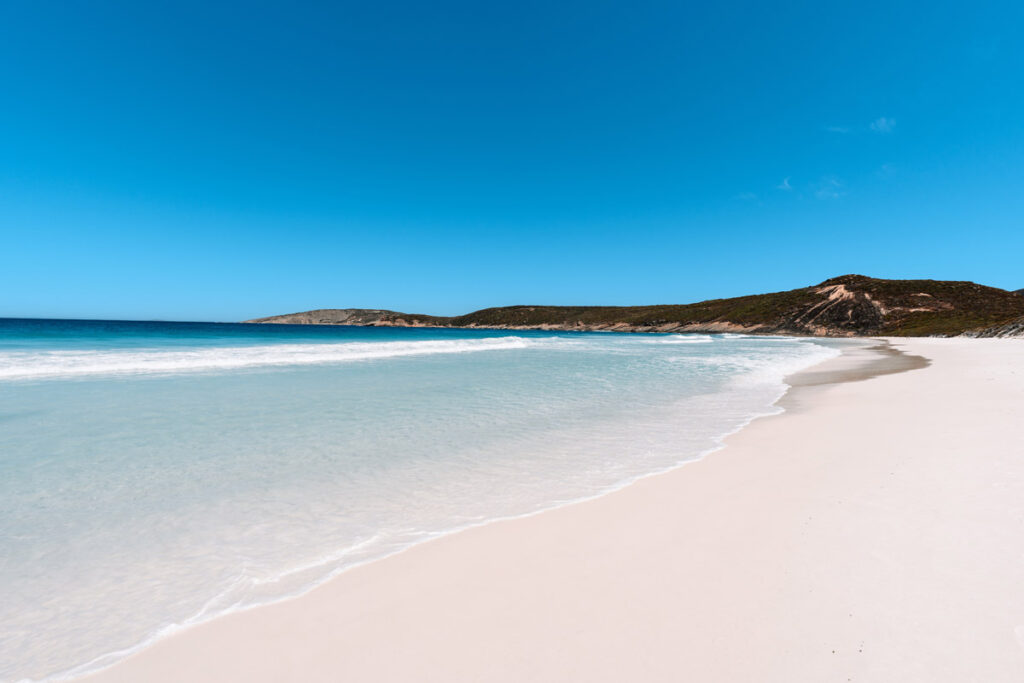 Minimalistische Aufnahme der Wasserlinie am Hellfire Bay. Das extrem klare Meerwasser läuft sanft auf den schneeweißen Sandstrand auf, unter einem wolkenlosen blauen Himmel.