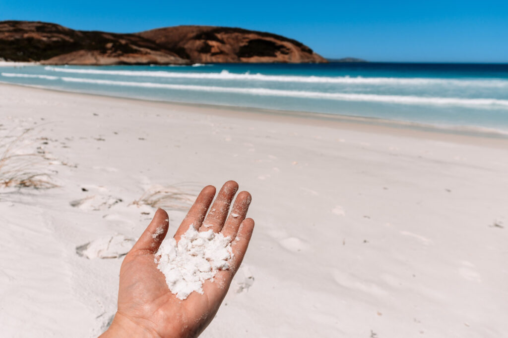 Nahaufnahme von Danis Hand, die feinen, weißen Quarzsand vom Hellfire Bay Strand hält. Im unscharfen Hintergrund sieht man das blaue Meer und die Bucht. Das Bild verdeutlicht die feine Qualität des Sandes.