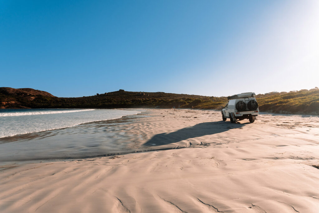 Ein weißer Geländewagen (4WD) fährt bei tiefstehender Sonne über den festen, weißen Sand am Strand von Lucky Bay. Die Reifenspuren im Sand und das warme Licht erzeugen eine abenteuerliche Roadtrip-Atmosphäre.