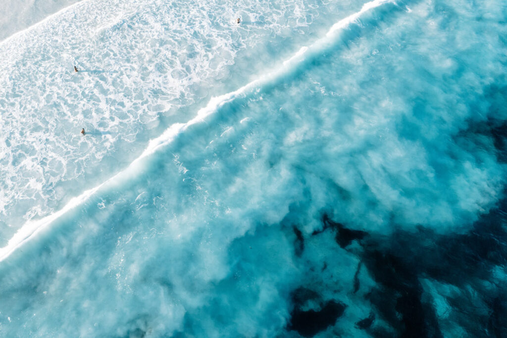 Detailaufnahme (Top-Down) der Meeresoberfläche in Lucky Bay. Weißer Schaum einer brechenden Welle bildet Muster auf dem intensiv türkisfarbenen Wasser, das die Reinheit des Ozeans hier verdeutlicht.