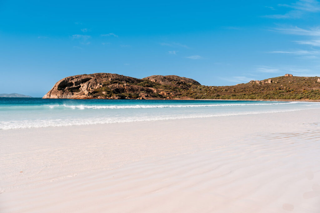 Traumhafte Aussicht auf die menschenleere Lucky Bay. Der weiße Sandstrand erstreckt sich weitläufig vor dem türkisfarbenen Wasser, im Hintergrund begrenzt ein grüner Hügel die Bucht.