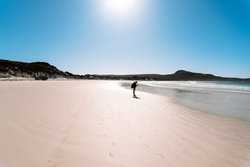 Weitwinkelaufnahme des riesigen Lucky Bay Strandes bei strahlendem Sonnenschein. Reiseblogger Mario steht klein am Wasser und fotografiert die Landschaft, was die gewaltigen Dimensionen der Bucht verdeutlicht.