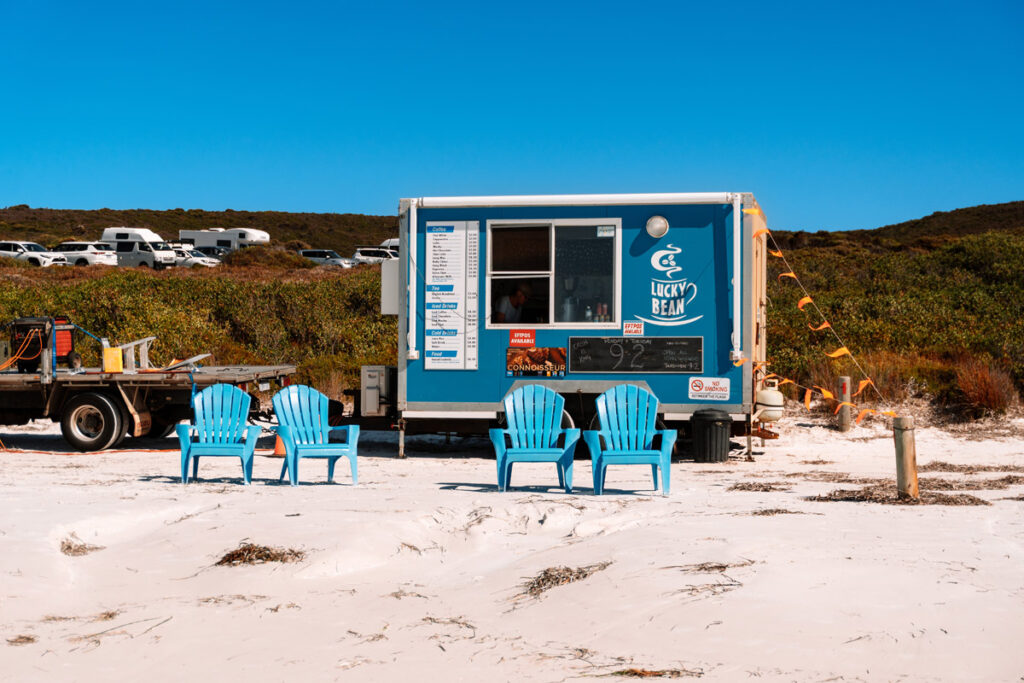 Der berühmte blaue 'Lucky Bean' Kaffeewagen (Food Truck) steht direkt am Strand von Lucky Bay. Davor laden blaue Stühle Gäste ein, ihren Kaffee mit Meerblick zu genießen.