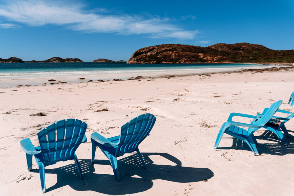Vier blaue Kunststoff-Gartenstühle stehen im weißen Sand am Lucky Bay Strand und laden zum Entspannen ein. Im Hintergrund leuchtet das türkisfarbene Meer.