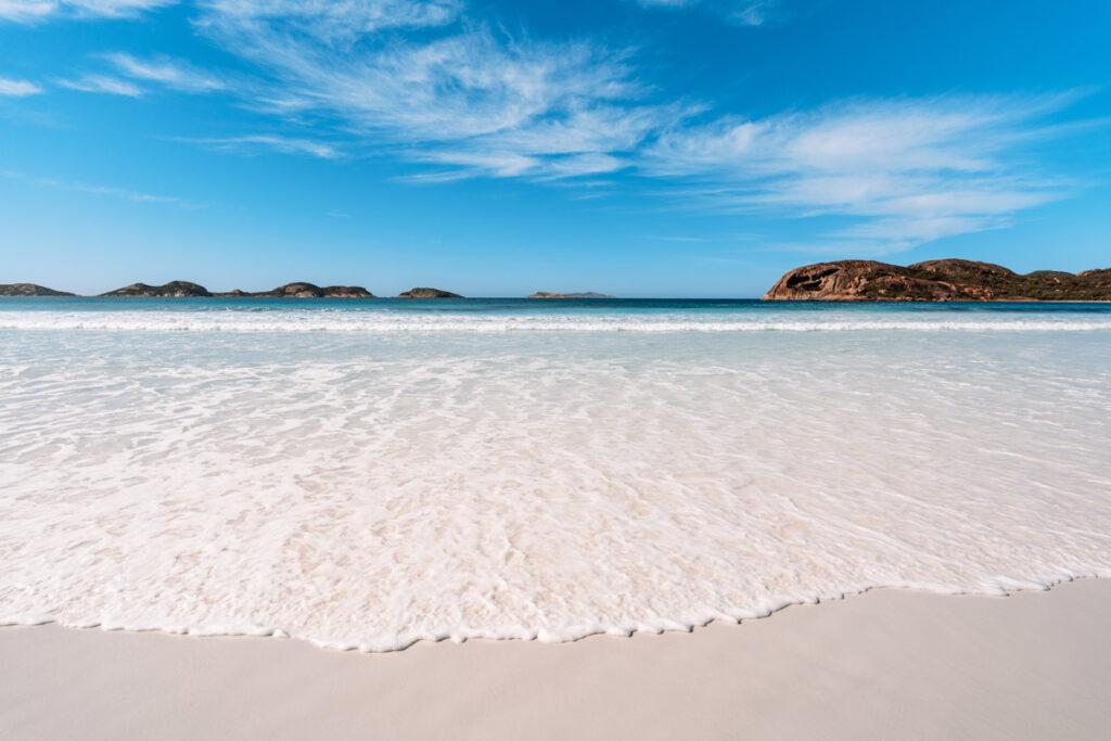 Ruhiges, flaches Wasser am Ufer der Lucky Bay. Die kleinen Wellen kräuseln sich sanft über den weißen Sandboden, darüber spannt sich ein blauer Himmel mit leichten Schleierwolken.