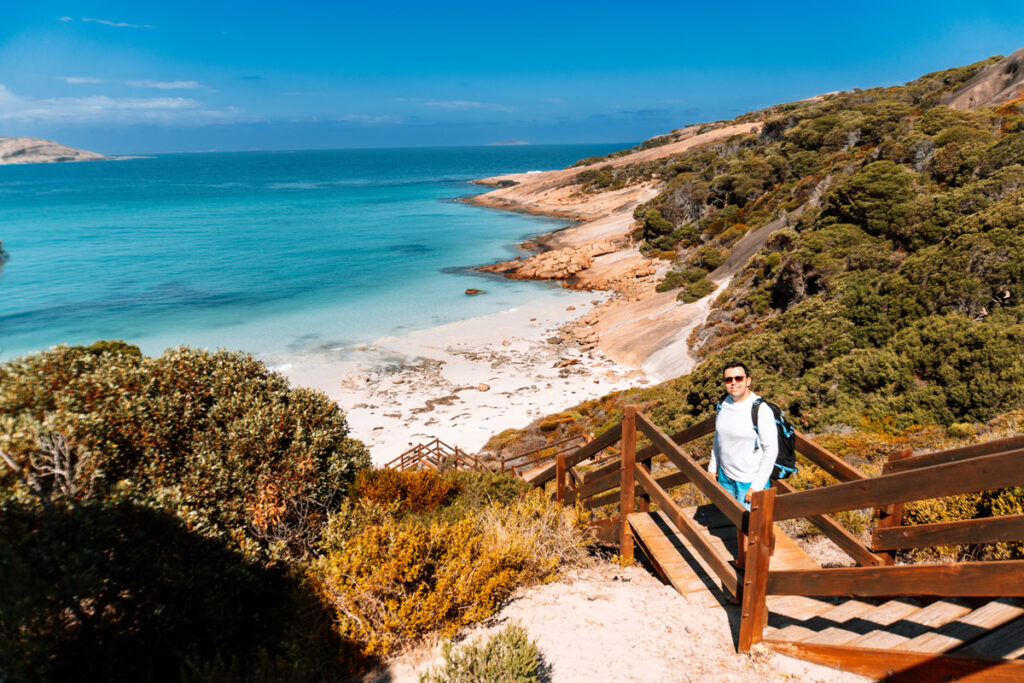 Reiseblogger (Mario von Auszeit Abenteuer) steht auf der steilen Holztreppe, die zum Blue Haven Beach hinunterführt. Er trägt Sonnenbrille und einen Rucksack, im Hintergrund leuchtet das unglaublich klare, türkisfarbene Wasser der Bucht. Die Küste ist von rötlichen Felsen und dichter, grüner Vegetation gesäumt.