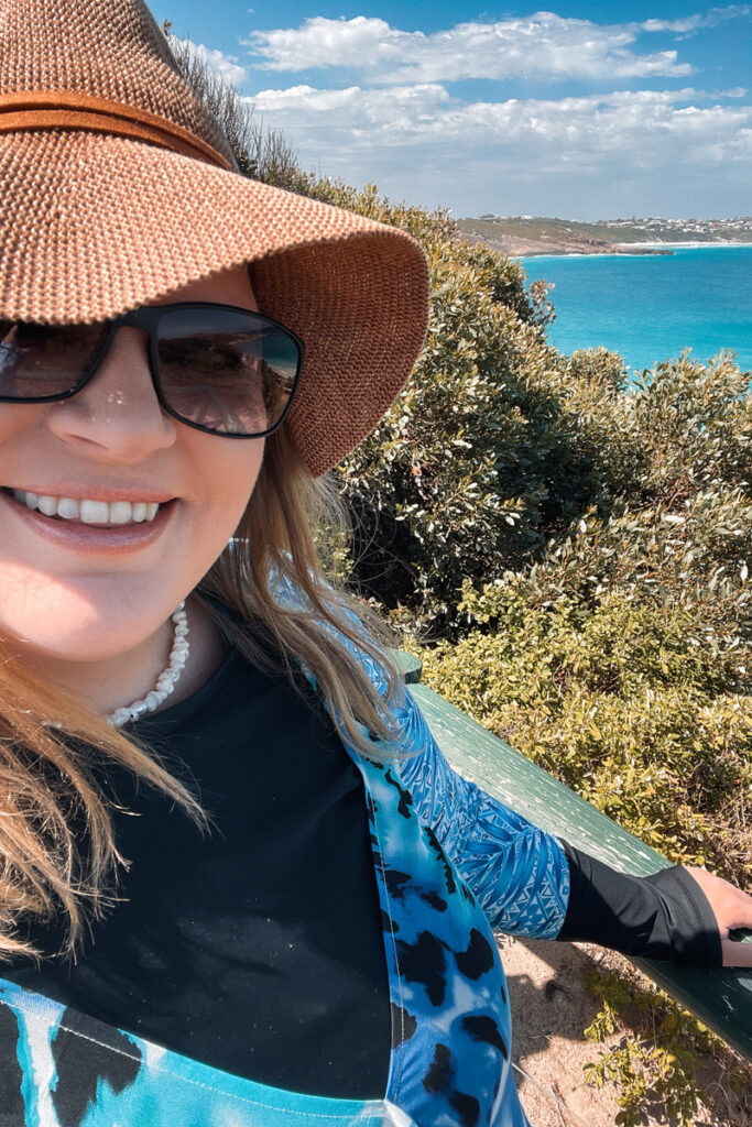Dani von Auszeit Abenteuer lächelt in die Kamera am Aussichtspunkt Blue Haven. Sie trägt einen Sonnenhut, Sonnenbrille, Schnorchelshirt und ein Sommerkleid darüber. Im Hintergrund sieht man das leuchtend türkise Wasser und grüne Sträucher.