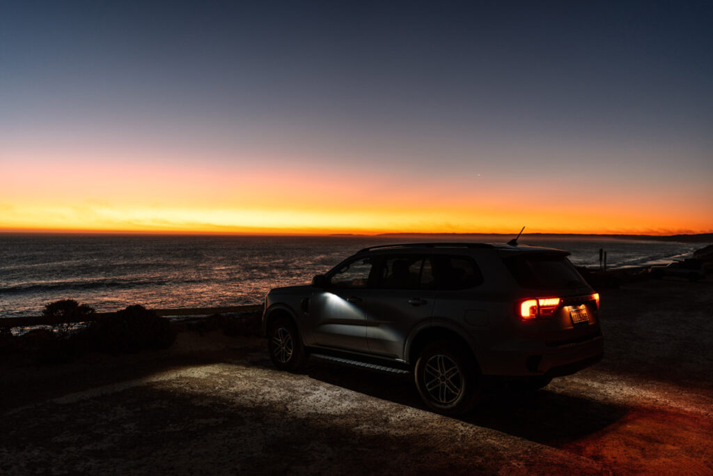 Ein Geländewagen (SUV) steht in der Abenddämmerung an der Küste am Nine Mile Beach. Der Himmel zeigt einen beeindruckenden Farbverlauf von Dunkelblau zu leuchtendem Orange am Horizont, wo die Sonne bereits untergegangen ist. Die Rücklichter des Autos leuchten rot, und die Scheinwerfer werfen Licht auf den sandigen Boden. Im Hintergrund sieht man das dunkle Meer.