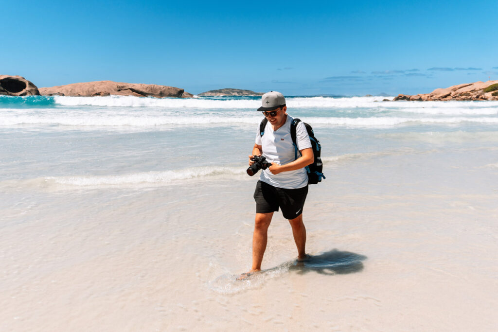 Reiseblogger (Mario, Auszeit Abenteuer) läuft barfuß durch das flache, klare Wasser am Twilight Beach. Er trägt eine Kamera in der Hand, einen Rucksack und eine Cape, während er den weißen Sandstrand erkundet. Im Hintergrund sieht man sanfte Wellen und die typischen Felsen.