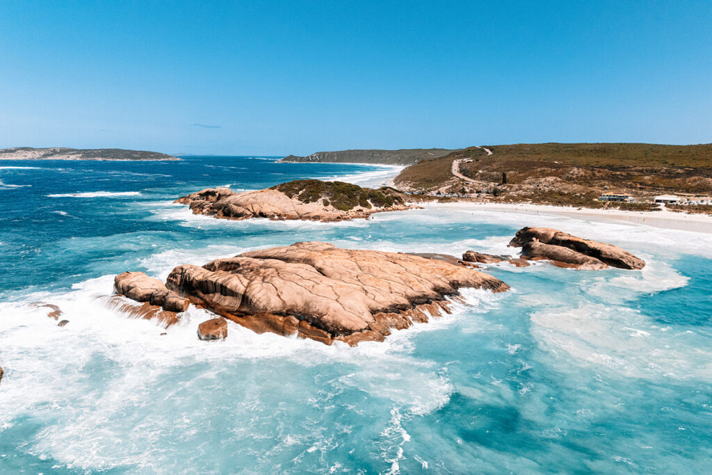 Beeindruckende Drohnenaufnahme von der Felseninsel im Meer vor dem Twilight Beach, Westaustralien. Das Bild zeigt den großen, braunen Granitfelsen im Vordergrund, der vom weiß schäumenden, türkisfarbenen Wasser umspült wird. Im Hintergrund erstreckt sich die lange Küstenlinie mit dem weißen Sandstrand, grünen Hügeln und einer Straße. Das Meer leuchtet in vielen Blau-Nuancen bis zum Horizont.