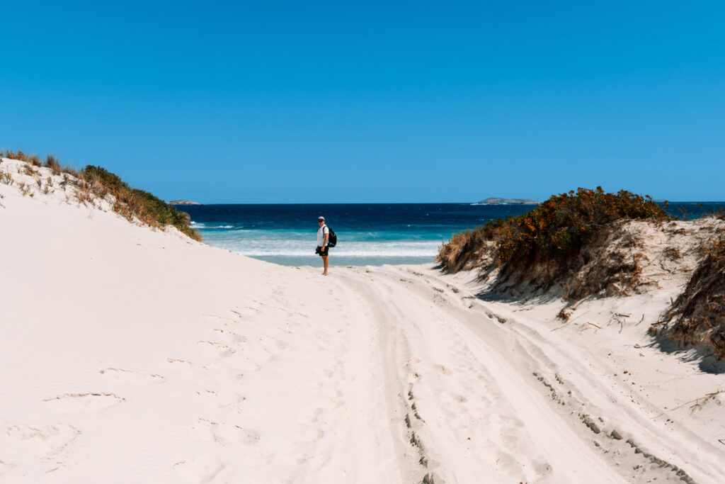Eine sandige Piste führt durch weiße Dünen zum Strand am Wylie Head. Im Hintergrund sieht man bereits das tiefblaue Meer und Mario von Auszeit Abenteuer, der den Weg erkundet. Der weiße Sand und der strahlend blaue Himmel vermitteln sofort Urlaubsstimmung.