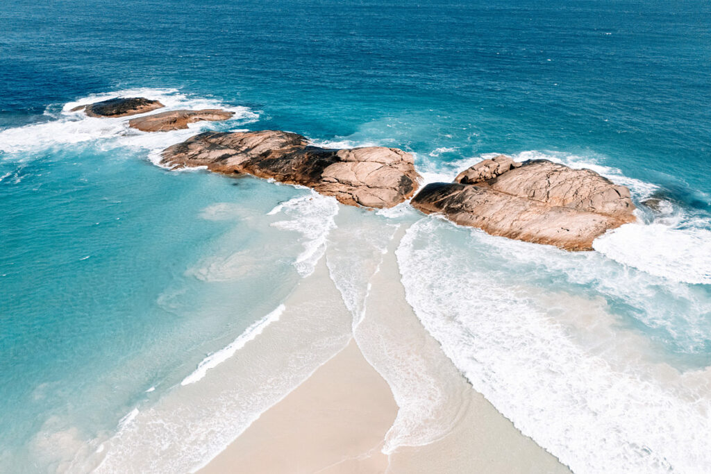 Spektakuläre Luftaufnahme von Felsenriffen im türkisfarbenen Wasser vor Wylie Head. Die Wellen umspülen die rötlichen Granitfelsen und bilden weiße Schaumkronen. Die Sandbank unter Wasser erzeugt wunderschöne Farbtöne von Hellblau bis Türkis.