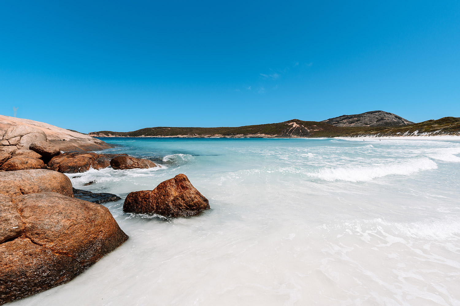 Traumhaftes Titelbild der Hellfire Bay im Cape Le Grand National Park, Westaustralien, aufgenommen von Auszeit Abenteuer. Das kristallklare, türkisfarbene Wasser trifft auf rötliche Granitfelsen im Vordergrund und geht in einen strahlend weißen Sandstrand über. Der Himmel ist wolkenlos blau. Ein Reisebericht über Danis Lieblingsstrand.