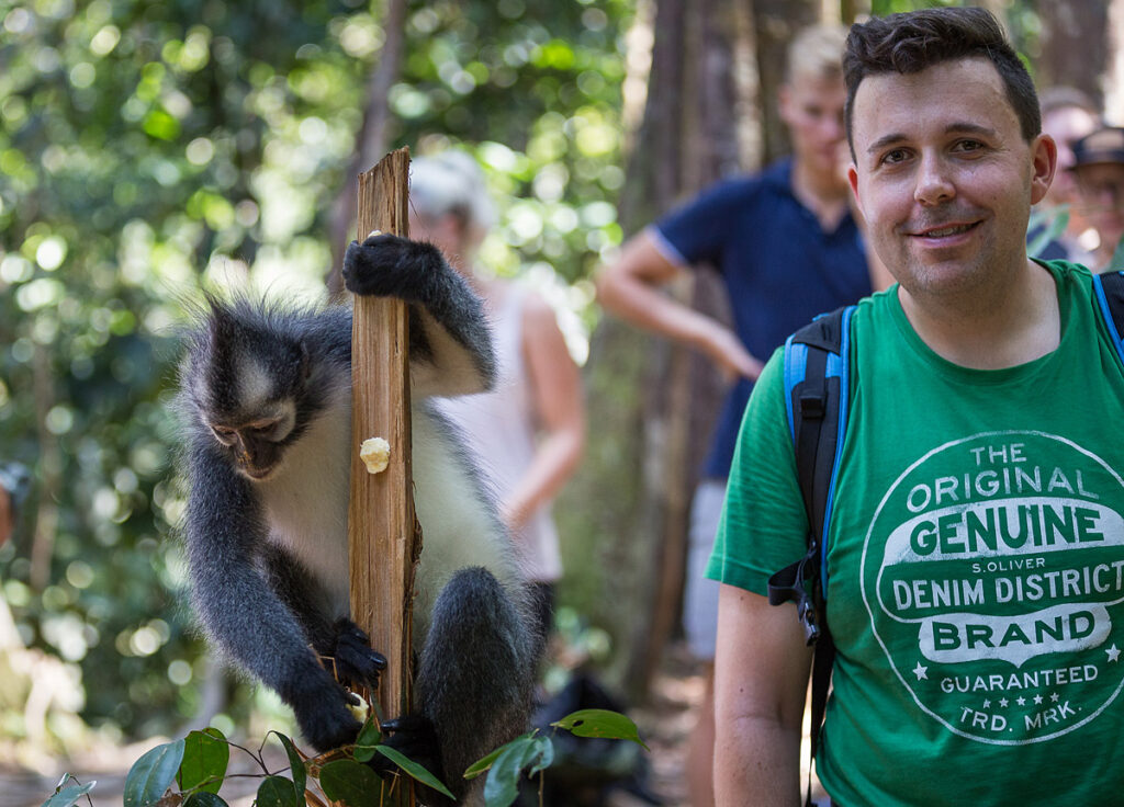 Mario von Auszeit Abenteuer lächelt in die Kamera, während ein Thomas-Langur neben ihm nach Obst an einem Holzpfosten greift Gunung Leuser Nationalpark Bukit Lawang Dschungeltrekking