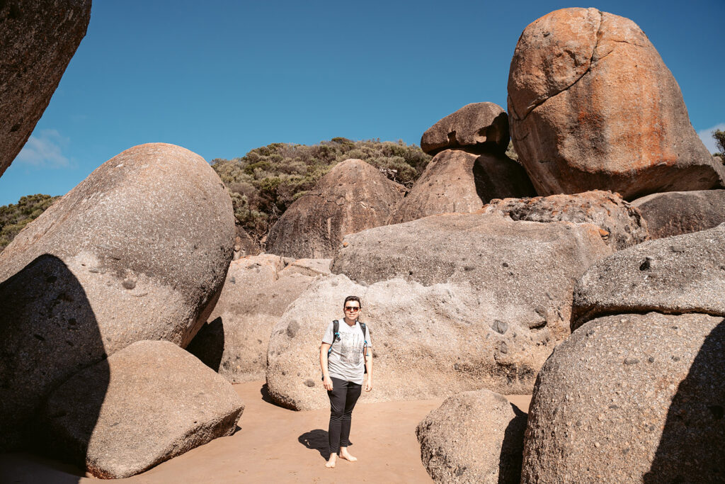 Mario vom Auszeit Abenteuer Reiseblog steht barfuß auf dem Sand vor massiven Granitfelsen an der Whisky Bay im Wilsons Promontory Nationalpark, Australien. Er trägt eine Sonnenbrille und ein helles T-Shirt. Der Hintergrund zeigt dichtes Küstengestrüpp unter einem blauen Himmel.