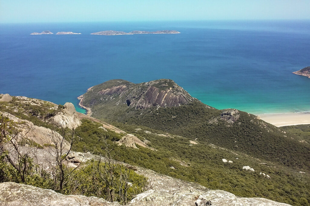 Blick über massive, graue Granitfelsen vom Gipfel des Mount Oberon im Wilsons Promontory Nationalpark. Im Hintergrund erstreckt sich die weite Küste mit dem Norman Beach und dem Ozean unter wolkenlosem Himmel.