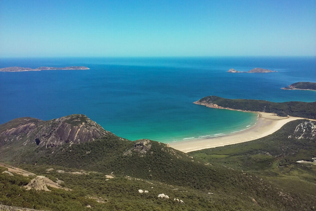 Atemberaubende Panoramasicht vom Gipfel des Mount Oberon im Wilsons Promontory Nationalpark. Das Bild zeigt den weiten Bogen des Norman Beach mit hellem Sand und türkisfarbenem Ozean. Im Hintergrund liegen felsige Inseln im tiefblauen Wasser unter strahlendem Himmel.