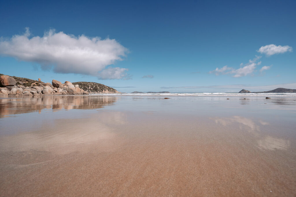 Weite Strandlandschaft an der Whisky Bay im Wilsons Promontory Nationalpark bei Ebbe. Der nasse, glatte Sand reflektiert den blauen Himmel mit Wolken und die ikonischen, rötlichen Granitfelsen auf der linken Seite. Ozeanwellen rollen sanft an Land in Australien.