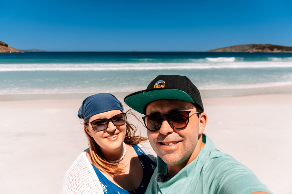 dani und mario vom team auszeit abenteuer lächeln in die kamera. beide tragen sonnenbrillen. hinter ihnen weißer sandstrand und türkises meer. im hintergrund sieht man links und rechts felsen im wasser. strand in westaustralien.