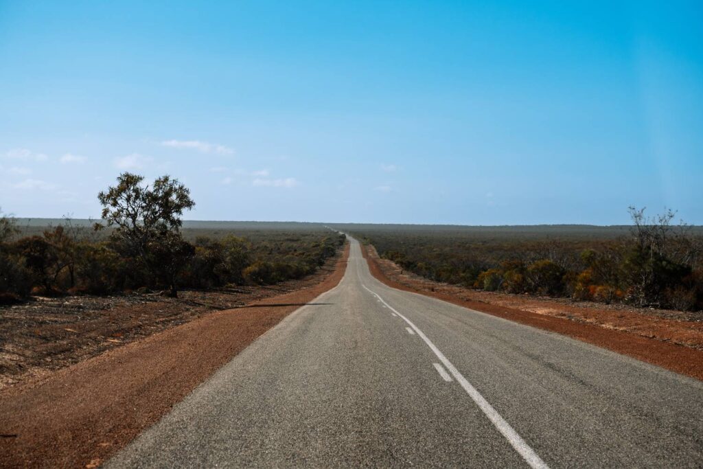 Eine endlose, asphaltierte Straße führt schnurgerade durch die karge, rötliche Outback-Landschaft Australiens unter einem klaren blauen Himmel.