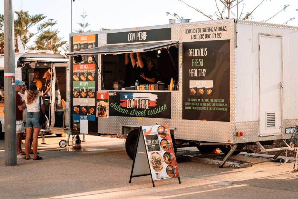 Ein silberner Foodtruck namens 'Con Pebre' bietet chilenische Street-Cuisine im warmen Abendlicht auf den Sunset Markets in Scarborough, Perth, während sich erste Gäste anstellen.