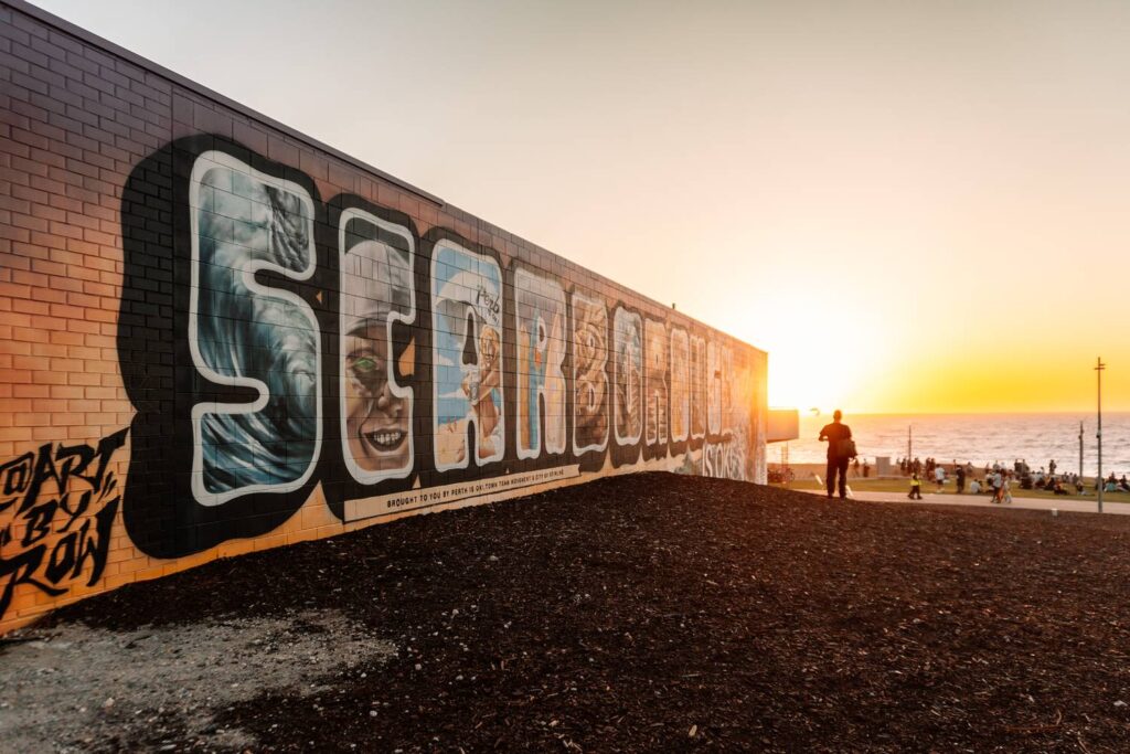 Ein großes, farbenfrohes Graffiti-Wandbild mit dem Schriftzug 'Scarborough' auf einer Mauer im warmen, goldenen Licht des Sonnenuntergangs an der Küste von Perth.