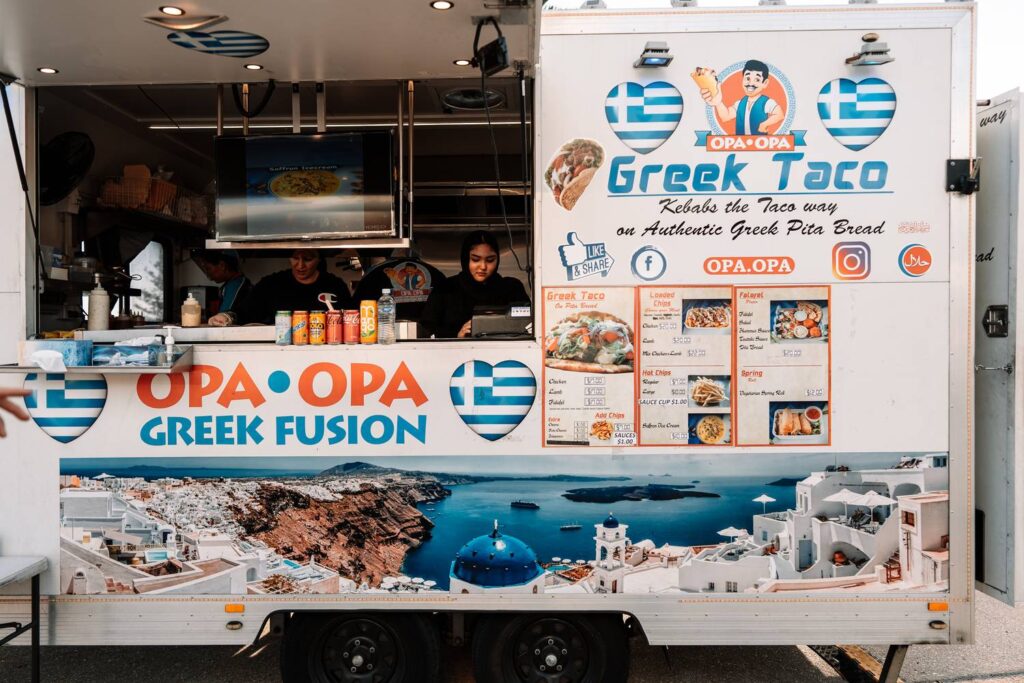 Der weiße Foodtruck 'Opa Opa Greek Fusion' steht verkaufsbereit beim Scarborough Sunset Market in Perth, dekoriert mit herzförmigen griechischen Flaggen, Menükarten und einer malerischen Santorini-Landschaft.