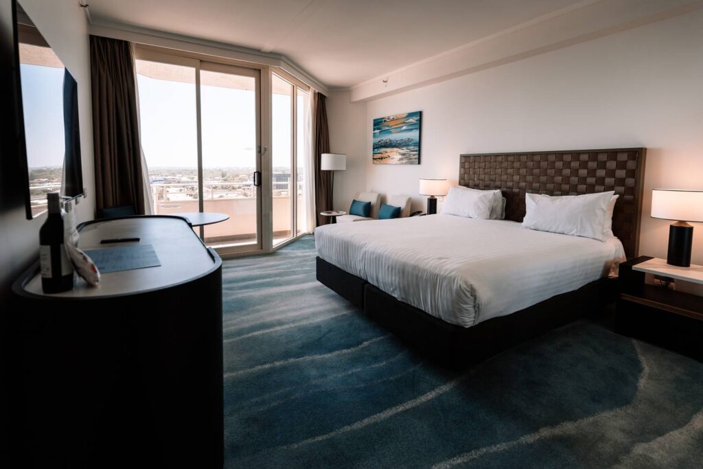 Ein stilvoll eingerichtetes Hotelzimmer in Scarborough mit einem großen Doppelbett, einem blauen Teppich in Wellenoptik und einer großen Fensterfront, die auf einen Balkon mit Weitblick führt.