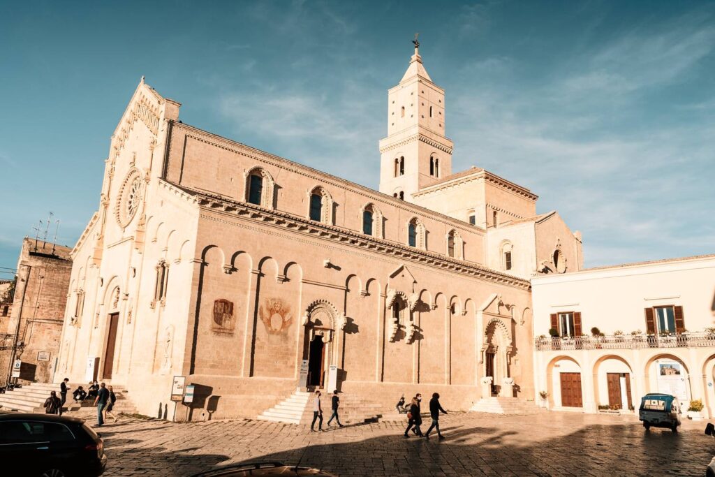 Die prachtvolle, helle Kathedrale von Matera ragt unter einem strahlend blauen Himmel empor, während Passanten den gepflasterten Vorplatz im warmen Nachmittagslicht überqueren.