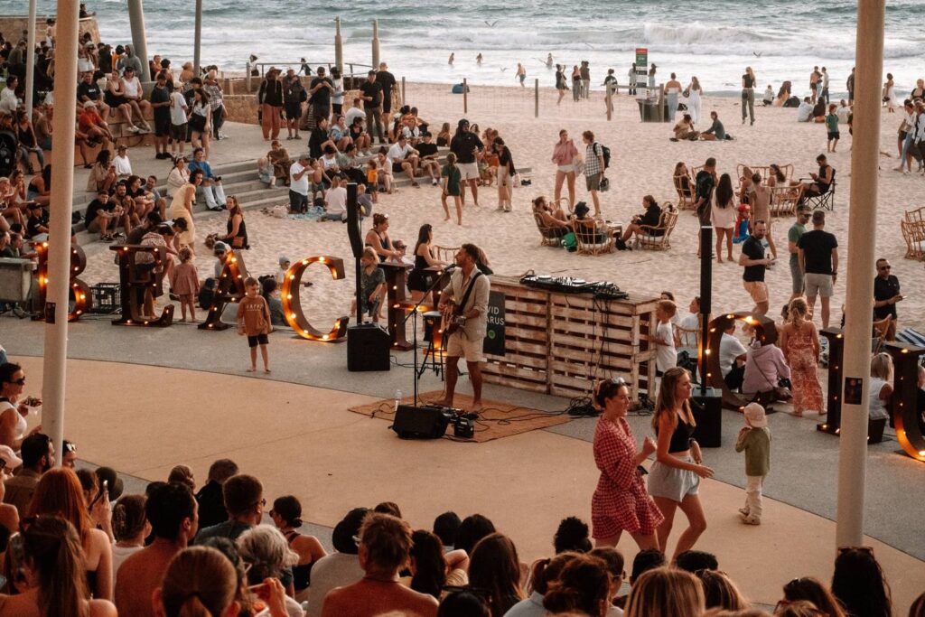David Lazarus mit Akustikgitarre tritt vor zahlreichen Zuschauern auf einer belebten Strandpromenade während der Sunset Markets am Scarborough Beach in Perth auf, während im Hintergrund die Wellen des Meeres brechen.