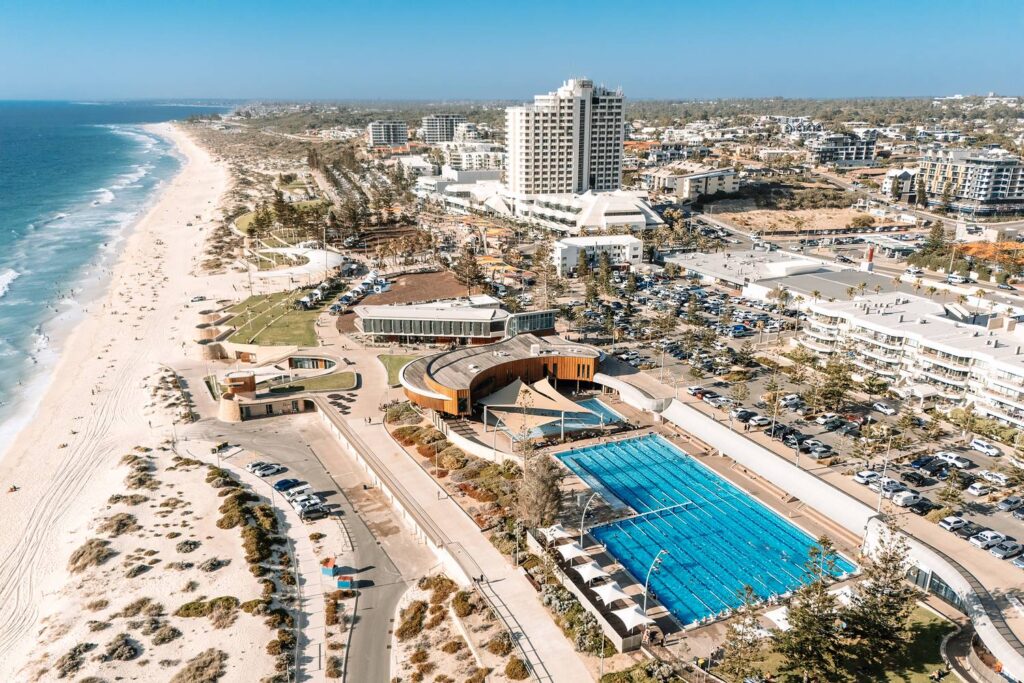 Eine sonnige Luftaufnahme des Scarborough Beach in Perth mit dem markanten, hellblauen Außenpool direkt am weißen Sandstrand und den angrenzenden Küstengebäuden unter strahlend blauem Himmel.
