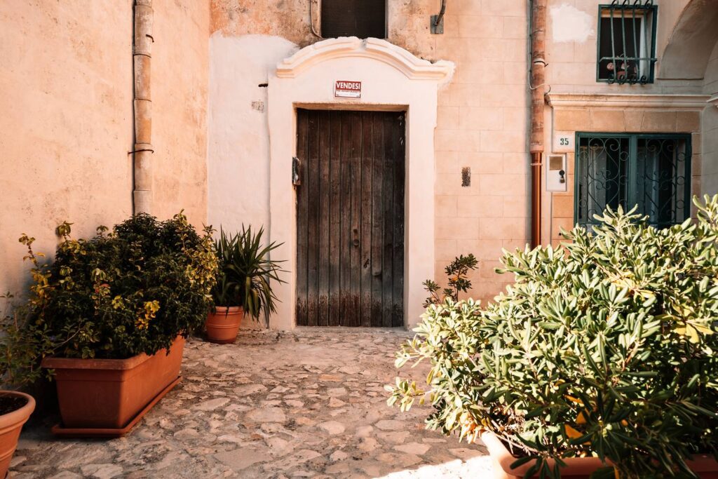 Idyllischer Fotospot in Matera: Eine alte Holztür umringt von zahlreichen Terrakotta-Blumentöpfen auf hellen Steinstufen.