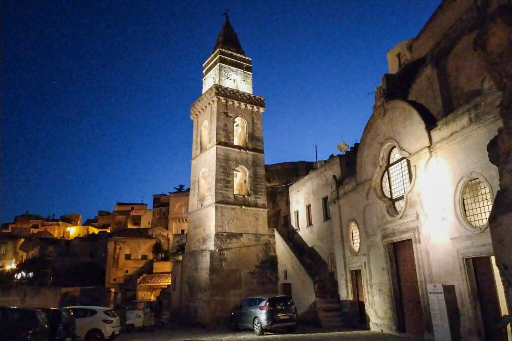 Der markante, warm beleuchtete Kirchturm der Kirche San Pietro Barisano ragt in der Altstadt Sassi von Matera vor einem tiefblauen Nachthimmel empor.