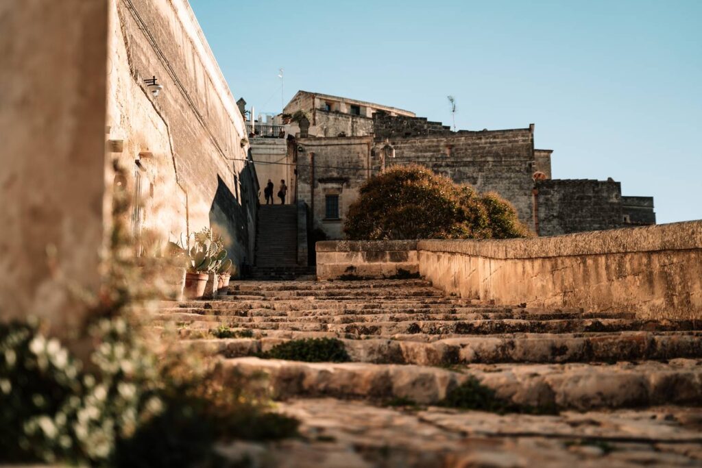 Blick über eine breite Steintreppe hinauf in die historische Altstadt Sassi von Matera, während Daniela und Mario im warmen Abendlicht zwischen den alten Steingebäuden stehen.