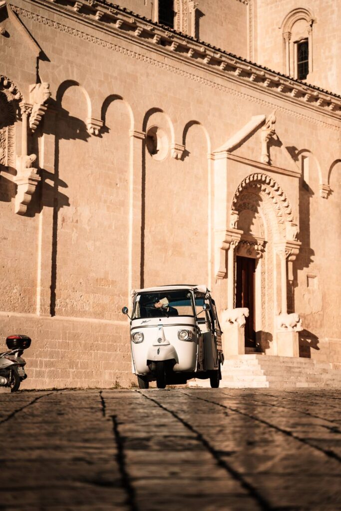 Ein klassisches, weißes Ape-Taxi parkt auf dem Kopfsteinpflaster vor einer historischen Steinfassade in Matera.