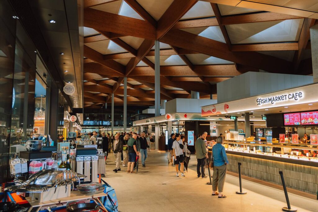 Besucher schlendern durch die lichtdurchflutete Halle des neuen Sydney Fish Market unter einer beeindruckenden, geometrischen Holzdeckenkonstruktion.