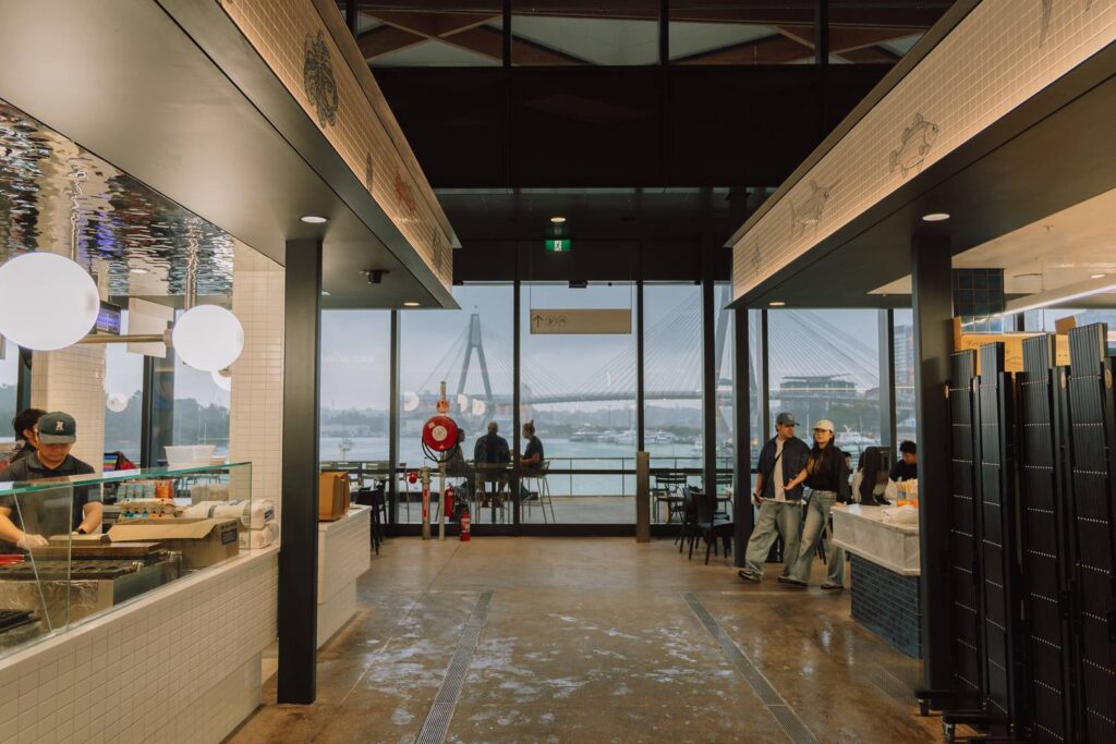 Moderner Innenbereich des New Sydney Fish Market mit Blick durch eine große Glasfront auf die Anzac Bridge und das Hafenbecken.