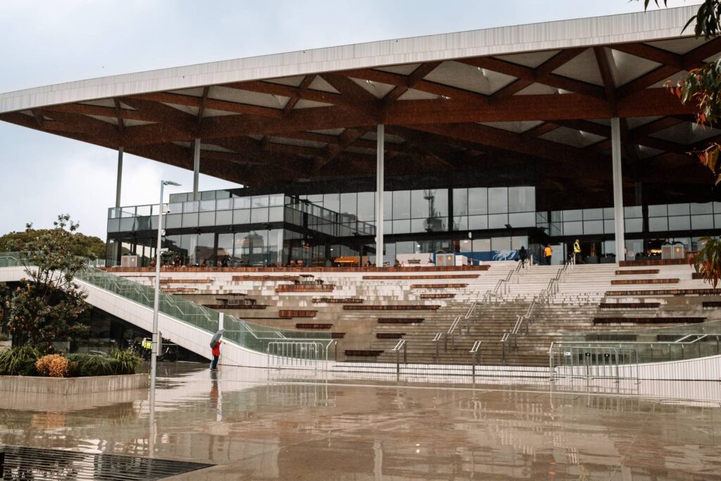 Die moderne Architektur des New Sydney Fish Market mit seinem markanten hölzernen Gitterdach und den weitläufigen Treppenaufgängen spiegelt sich auf dem nassen Vorplatz an einem regnerischen Tag.