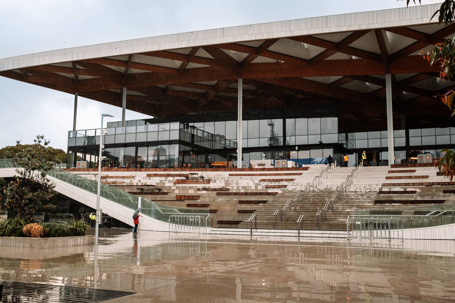 Die moderne Architektur des New Sydney Fish Market mit seinem markanten hölzernen Gitterdach und den weitläufigen Treppenaufgängen spiegelt sich auf dem nassen Vorplatz an einem regnerischen Tag.